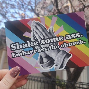 May include: A rectangular postcard with a rainbow background and the text "Shake some ass. Embarrass the church." The image features a black and white illustration of praying hands.