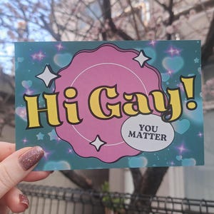 May include: A postcard with the text "Hi Gay!" in yellow bubble letters with a black outline, set against a pink background. The card has a teal background with stars and hearts. The text "You Matter" is in a white oval.