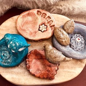 Peut inclure: Un ensemble de quatre petits vide-poches en argile sculptés à la main. Il y a un chat bleu sarcelle, un disque cuivré avec le mot DREAM et une fleur, un oiseau brun sur une feuille cuivrée, et un plat gris avec des feuilles et un cœur.