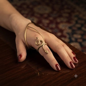 Puede incluir: Un adorno de mano de metal en tono dorado que presenta una banda delgada y curva que envuelve el dorso de la mano y se conecta a un anillo de serpiente texturizado y enroscado que rodea el dedo medio con un patrón de escamas detallado.