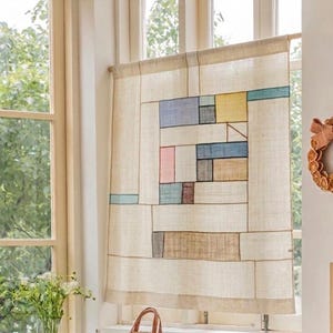 May include: A rectangular beige fabric curtain featuring a geometric patchwork design with thin dark brown lines. The pattern includes various rectangular blocks in muted shades of blue, yellow, pink, brown, and grey, suspended from a wooden rod.
