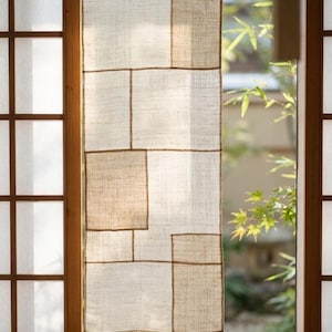 Puede incluir: Una cortina rectangular de lino beige con una cuadrícula de paneles rectangulares cosidos de varios tamaños. La tela tiene una textura tejida visible y es semitranslúcida, lo que permite que la luz pase a través del material al colgar.