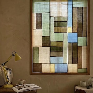 Puede incluir: Una cortina de ventana rectangular con un diseño de patchwork de paneles de tela translúcida en tonos verde salvia, marrón, crema y azul claro, enmarcada por un borde de madera e iluminada por la luz natural que filtra el tejido.