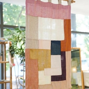 Puede incluir: Un tapiz rectangular compuesto por paneles de tela cosidos en varios colores, incluyendo rosa, blanco, beige, amarillo mostaza, azul oscuro, naranja óxido y morado intenso. La pieza textil cuelga de una barra de madera mediante trabillas.