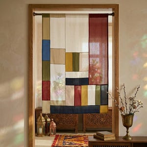 May include: A rectangular fabric curtain featuring a patchwork design of various solid colour blocks including cream, deep red, navy blue, olive green, and mustard yellow, suspended from a dark metal rod within a wooden door frame.