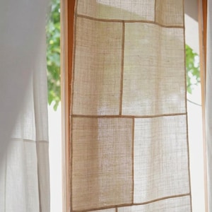 Puede incluir: Un panel de cortina beige con un diseño de patchwork compuesto por secciones rectangulares de tela tejida. El material tiene un tejido de fibra natural visible y texturizado con costuras distintas que conectan cada pieza rectangular.