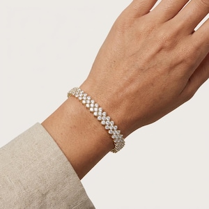 May include: A gold-toned bracelet with two rows of sparkling, clear gemstones. The bracelet is worn on a wrist, with a neutral-colored sleeve visible in the image. The gemstones are set closely together, creating a continuous, shimmering effect.