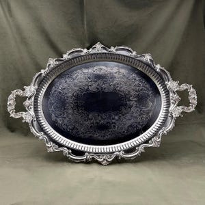 May include: An oval silver-toned serving tray featuring ornate, raised scrollwork along the scalloped edges and handles. The central surface displays a dark, polished finish with intricate, etched floral and scroll patterns throughout the interior.