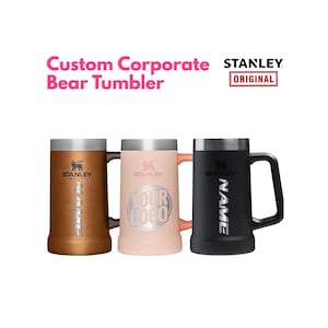 May include: Three Stanley brand beer steins with handles, featuring a metallic rim and base. The left stein is copper, the center is light pink with a circular logo, and the right is black. Each features the Stanley logo and the word NAME or YOUR LOGO.
