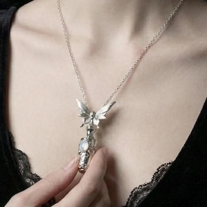 May include: Silver-toned necklace featuring a detailed butterfly pendant atop a small, ornate bottle with a round, iridescent stone. The necklace is worn against a dark background, highlighting the intricate design and delicate chain.