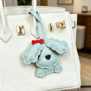 May include: A light blue leather dog-shaped bag charm with a red bow, hanging from a white leather handbag. The charm has a black nose and cloud-shaped ears. The handbag has gold-colored hardware.