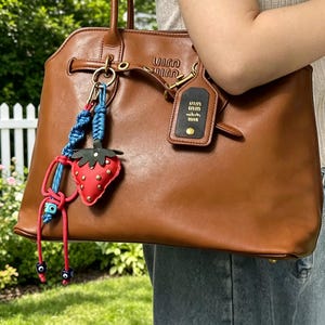 May include: A brown leather tote bag with a strawberry keychain and a black tag. The strawberry keychain is red with green leaves and blue and red accents. The black tag has white text and a gold rivet.