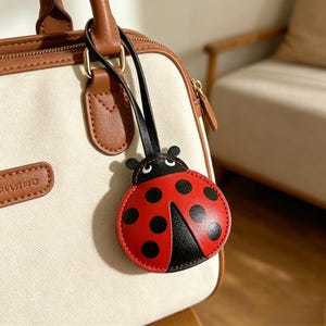 Puede incluir: Un colgante para bolso con forma de mariquita hecho de material rojo y negro, con una correa negra sujeta a un bolso de color crema y marrón. El colgante tiene un cuerpo redondeado con puntos negros, una cabeza negra y dos antenas.