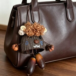 May include: A quirky brown bag charm depicting a tree-like character with curly brown hair, a button nose, and acorn details. The charm is suspended from a brown leather handbag, offering a playful and unique accessory.