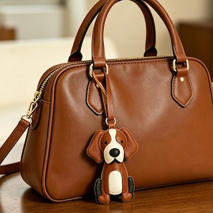 May include: A brown leather handbag featuring a top zip closure, two handles, and a detachable shoulder strap. A dog-shaped leather charm in brown, white, and black is attached to the front handle hardware, hanging against the bag's surface.