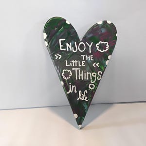 May include: A heart-shaped wooden sign with a dark green, purple, and white painted design. The sign features the words "Enjoy the little things in life" in white script, with decorative accents.
