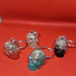 May include: Four resin skull keychains with silver key rings. The skulls have varying colors, including red, green, and black, with a clear, glittery finish. The keychains are set against a red background.