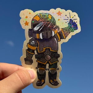May include: A holographic sticker featuring a stylized armored character in dark gray and yellow plating, wearing a helmet with a visor, giving a thumbs up. The design includes small orange star accents and a skull emblem on the shoulder.