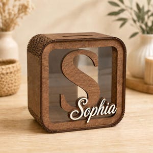 May include: A square wooden money box featuring a large cutout letter S on the front panel. A clear acrylic window is set behind the letter, and the name Sophia is written in white cursive script across the bottom right of the front frame.