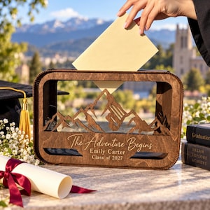 May include: A rectangular wooden card box with a laser-cut mountain range silhouette. The front is engraved with the text "The Adventure Begins," "Emily Carter," and "Class of 2027." A hand inserts a cream-colored envelope into the top slot.