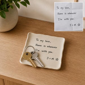 以下が含まれることがあります： オフホワイトの正方形のセラミック製トレイ。黒い文字で「To my love, Home is wherever I'm with you. J + M」と星のマークがプリントされています。シルバーのキーリングに通された2本の金属製の鍵がトレイの上に置かれています。