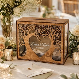 May include: A rectangular wooden card box with a laser-cut tree design forming a heart shape in the center. The clear front panel features the white printed text Emma & Noah and the date 06.21.2027. The box has a slot on top for inserting cards.