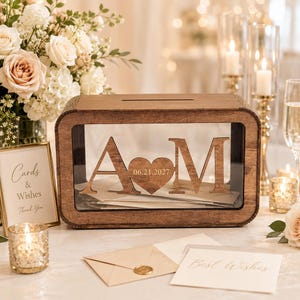 Puede incluir: Una caja de madera rectangular para tarjetas con un panel frontal transparente que muestra las letras recortadas A y M separadas por un corazón grabado con la fecha 06.21.2027. La caja tiene una ranura en la parte superior y acabado en madera.
