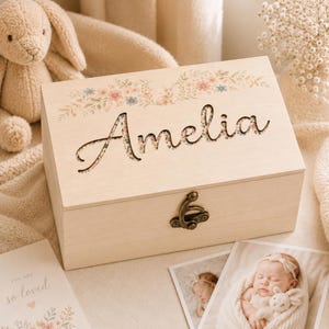 May include: A rectangular light-coloured wooden keepsake box featuring the name Amelia cut out in a cursive font. The lid is decorated with a floral and bunny illustration. A bronze-coloured metal latch secures the front of the wooden box.