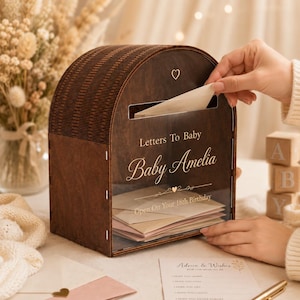 Könnte beinhalten: Eine dunkelbraune Holzbox mit gewölbtem Deckel und einer transparenten Vorderseite. Der Text lautet Letters To Baby Baby Amelia und Open On Your 18th Birthday. Ein kleines Herzsymbol ist über dem Schlitz eingraviert. Die Box enthält Briefumschläge.