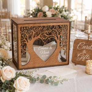 May include: A rectangular wooden card box with a laser-cut tree design forming a heart shape in the center. The front panel features the white printed text "Emma & Noah" and "06.21.2027". The box has clear side panels and a slot on top.