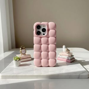 May include: A light pink phone case with a puffy, quilted design. The case has a square pattern and a camera cutout. The phone case is sitting on a white marble surface.