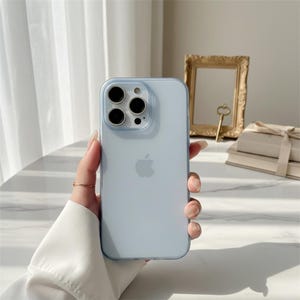 Könnte beinhalten: Eine hellblaue iPhone-Hülle mit transparentem Rand. Die Hülle hat eine glatte Oberfläche und zeigt das Apple-Logo. Die Handyhülle wird in einer Hand gehalten, mit einem goldenen Ring am Finger.