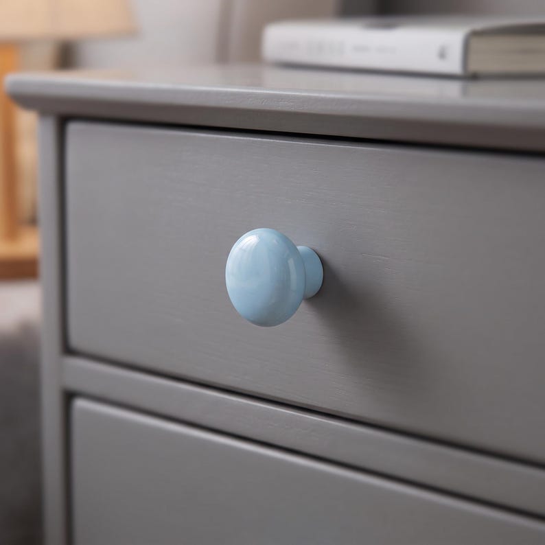 Peut inclure: Un bouton de meuble rond et lisse en c&eacute;ramique bleu clair fix&eacute; sur une fa&ccedil;ade de tiroir en bois gris. Le bouton pr&eacute;sente une finition brillante et une forme sph&eacute;rique simple, contrastant avec la surface grise mate du tiroir sur lequel il est mont&eacute;.