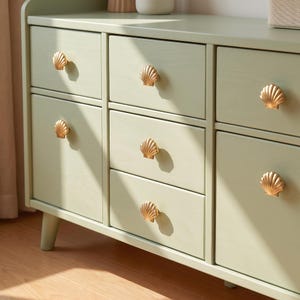 May include: A sage green wooden dresser featuring six drawers arranged in three columns. Each drawer is equipped with a single gold-toned metal knob shaped like a seashell. The furniture piece stands on short, tapered legs and has a smooth finish.