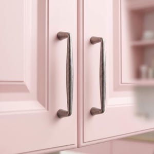 May include: Two vertical cabinet handles with a dark, weathered metallic finish attached to light pink cabinet doors. The handles feature a smooth, elongated, slightly curved design with rounded mounting points at the top and bottom ends.