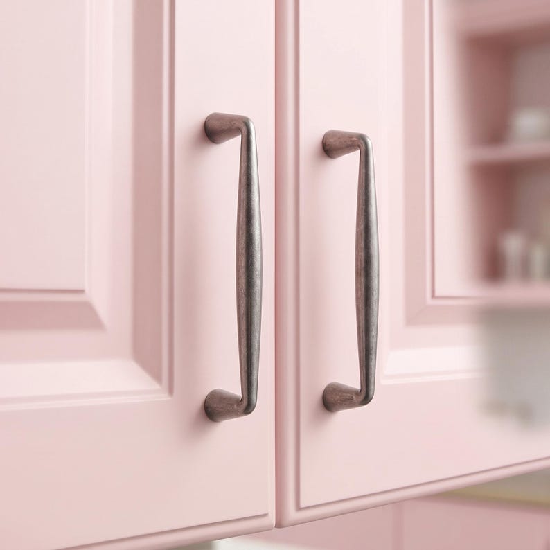 May include: Two vertical cabinet handles with a dark, weathered metallic finish attached to light pink cabinet doors. The handles feature a smooth, elongated, slightly curved design with rounded mounting points at the top and bottom ends.