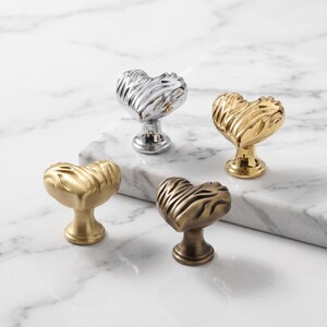 May include: Four heart-shaped cabinet knobs with a textured, rippled surface. The set includes one polished chrome knob, one shiny gold knob, one brushed brass knob, and one antique bronze knob, each featuring a central pedestal base.