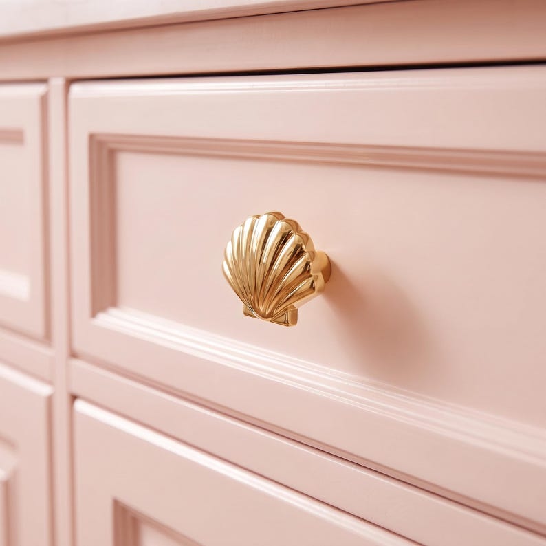 May include: A shiny gold-toned metal drawer knob shaped like a seashell with vertical ridges. The knob is mounted on a light pastel pink wooden drawer front featuring a rectangular recessed panel design and smooth, matte finish surfaces.