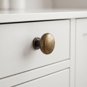 May include: A round cabinet knob with a brushed antique brass finish. The knob features a smooth, slightly domed surface and a circular base plate, mounted onto a flat, light-colored drawer front with horizontal paneling details.
