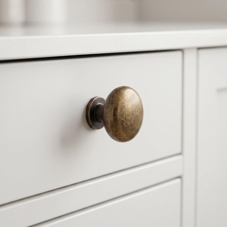May include: A round cabinet knob with a brushed antique brass finish. The knob features a smooth, slightly domed surface and a circular base plate, mounted onto a flat, light-colored drawer front with horizontal paneling details.