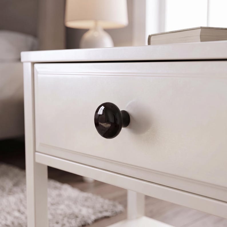 Peut inclure: Un bouton de meuble rond et noir brillant est centr&eacute; sur la fa&ccedil;ade d'un tiroir en bois blanc. Le bouton pr&eacute;sente une surface lisse et r&eacute;fl&eacute;chissante ainsi qu'une forme circulaire simple, cr&eacute;ant un contraste marqu&eacute; avec la finition blanc cass&eacute;.