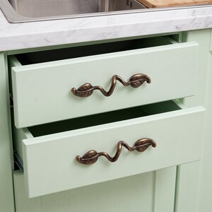 May include: Two light green kitchen drawers featuring antique bronze-colored metal pulls. Each handle has a winding, vine-like shape with two detailed leaf accents. The drawers are open, revealing the interior space against a white countertop.