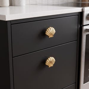 May include: Two gold-toned metal cabinet knobs shaped like seashells with vertical ridges. These decorative hardware pieces are mounted onto the front of two stacked matte black kitchen drawers, providing a metallic contrast to the dark surface.