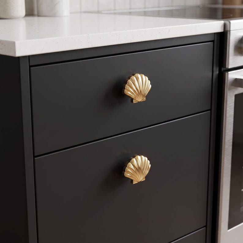 May include: Two gold-toned metal cabinet knobs shaped like seashells with vertical ridges. These decorative hardware pieces are mounted onto the front of two stacked matte black kitchen drawers, providing a metallic contrast to the dark surface.