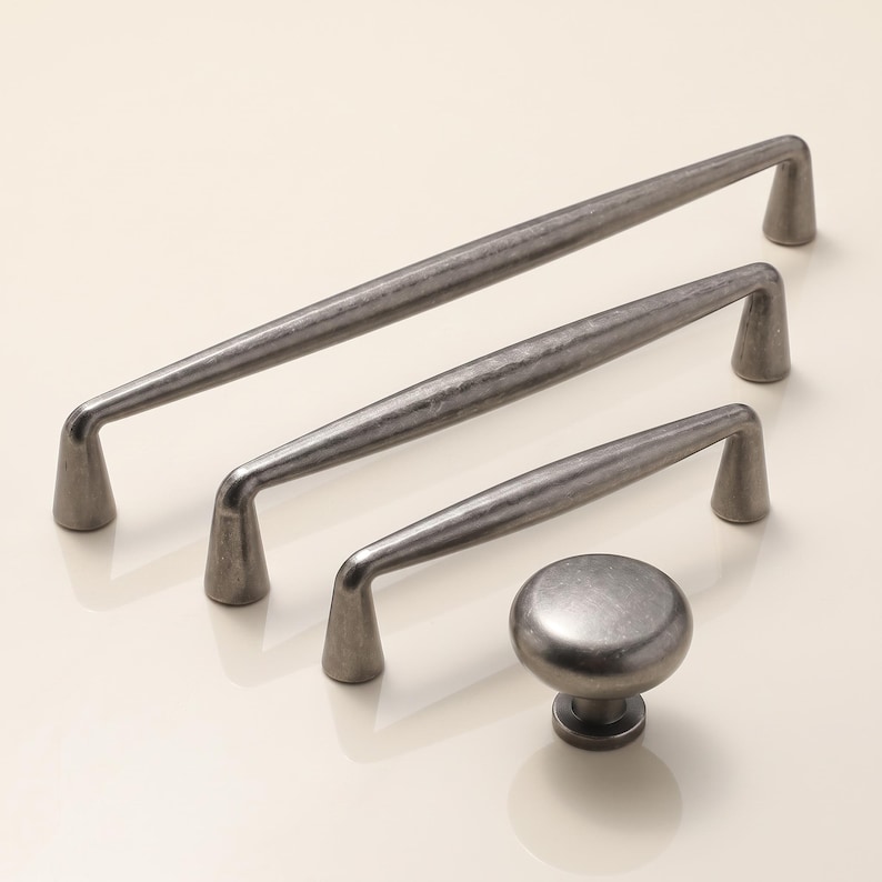 May include: A collection of four cabinet hardware pieces in a dark, brushed metallic finish. The set includes three elongated bar pulls of varying lengths with tapered feet and one circular knob with a matching textured, weathered appearance.