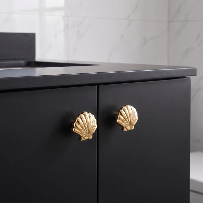 May include: Two gold-toned cabinet knobs shaped like seashells are mounted on a matte black cabinet door. The knobs feature a detailed, ridged fan-like texture that mimics the appearance of a natural scallop shell, providing a metallic contrast.
