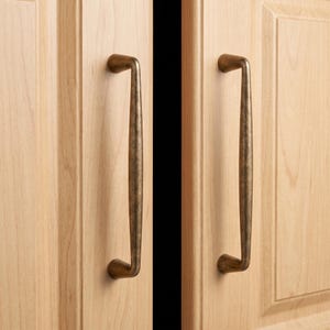 May include: Two vertical cabinet pulls with a dark, weathered bronze finish and a smooth, elongated cylindrical shape. Each handle features a slightly tapered design with rounded ends, mounted onto light-toned wooden cabinet doors with panels.