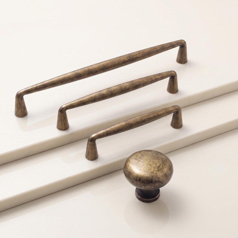 May include: A collection of cabinet hardware featuring three elongated metal pulls of varying lengths and one round metal knob. Each piece has a textured, antique brass finish with a dark, weathered patina and tapered mounting points.