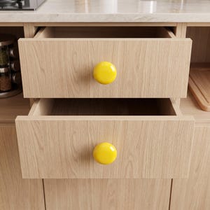 Peut inclure: Deux tiroirs de cuisine en bois clair &agrave; grain apparent, empil&eacute;s verticalement, chacun dot&eacute; d'un bouton circulaire unique, lisse, brillant et jaune vif, centr&eacute; sur la fa&ccedil;ade. Les tiroirs sont ouverts, r&eacute;v&eacute;lant la structure int&eacute;rieure du meuble.