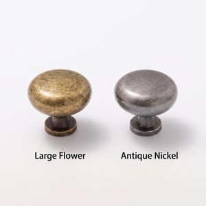 May include: Two round cabinet knobs with a smooth, domed top and a short cylindrical base. The left knob has a textured, mottled brass finish labeled Large Flower, and the right knob has a smooth, dark metallic finish labeled Antique Nickel.